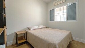 Quarto com cama, mesa de cabeceira, janela com persianas e ar condicionado.