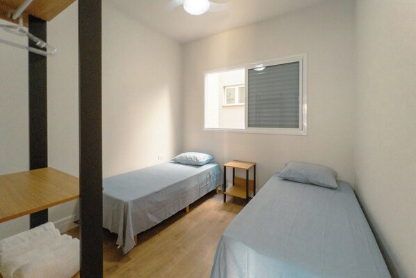 A small, single-room accommodation with two single beds, a wooden table, and a window with blinds.