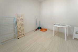 A room with a metal rack, a wooden ladder, a mop, a broom, and a table with a chair.