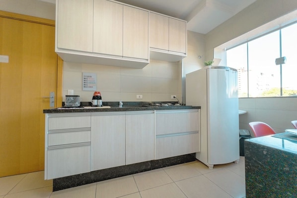 A kitchen with white cabinets, a black countertop, a white refrigerator, and a window providing natural light. - Zona 7 Apartment with AC and Wi-Fi - MN204 (Maringá)