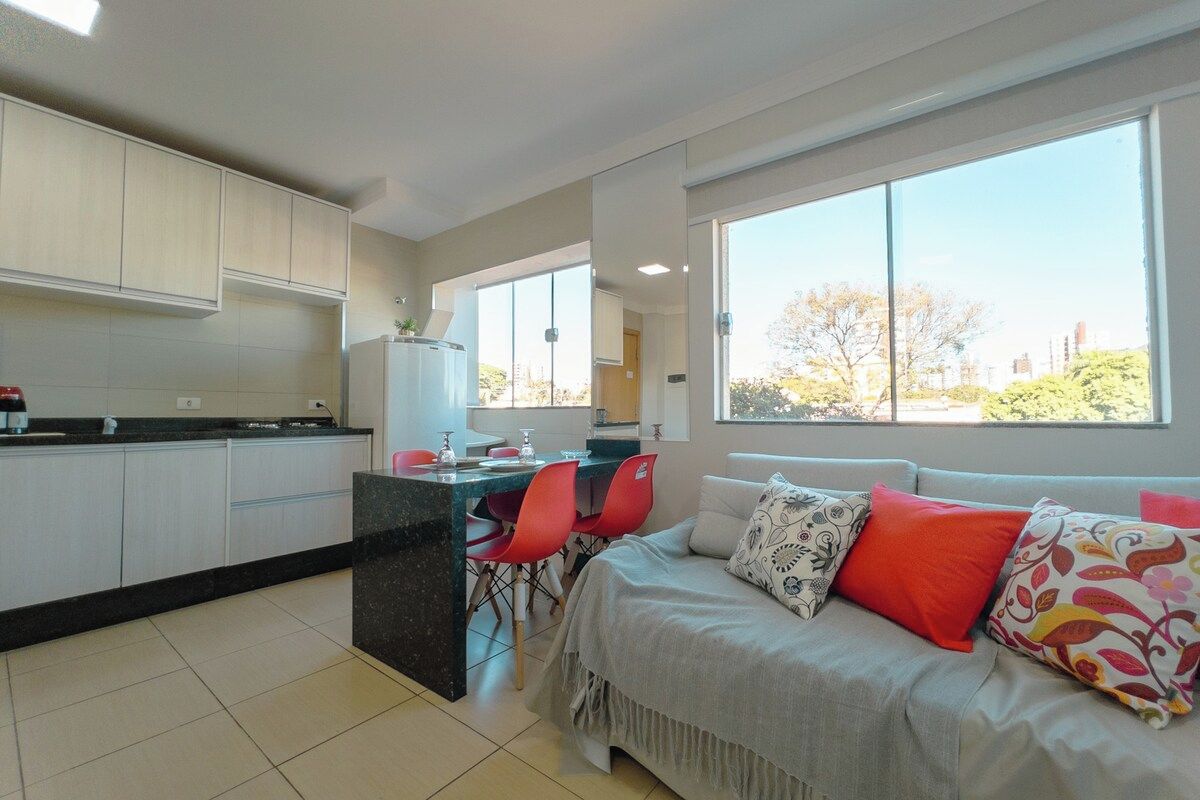 A modern kitchen with white cabinets, a dining area with red chairs, and a large window with a view of trees and buildings.