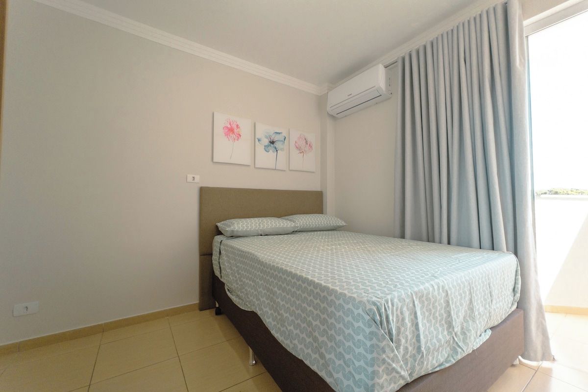 A bedroom with a bed, a headboard, a window with curtains, and a wall-mounted air conditioner.