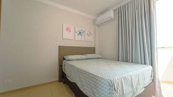 A bedroom with a bed, a headboard, a window with curtains, and a wall-mounted air conditioner.