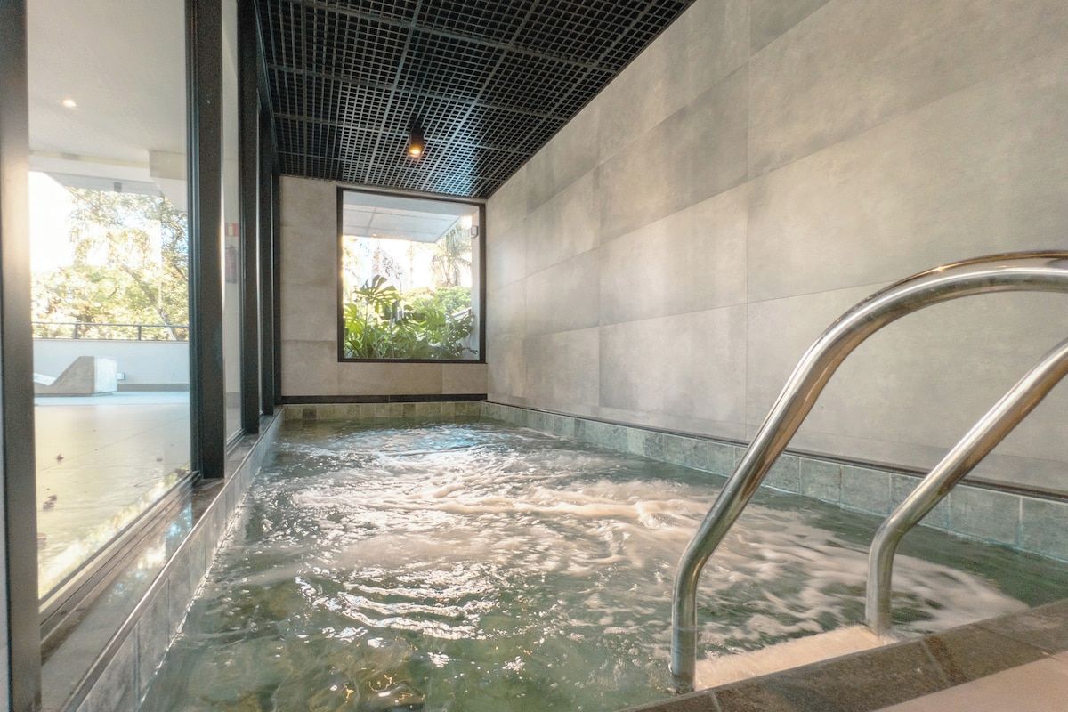 Un jacuzzi con escalera metálica, rodeado de un muro de azulejos y un ventanal de vidrio.