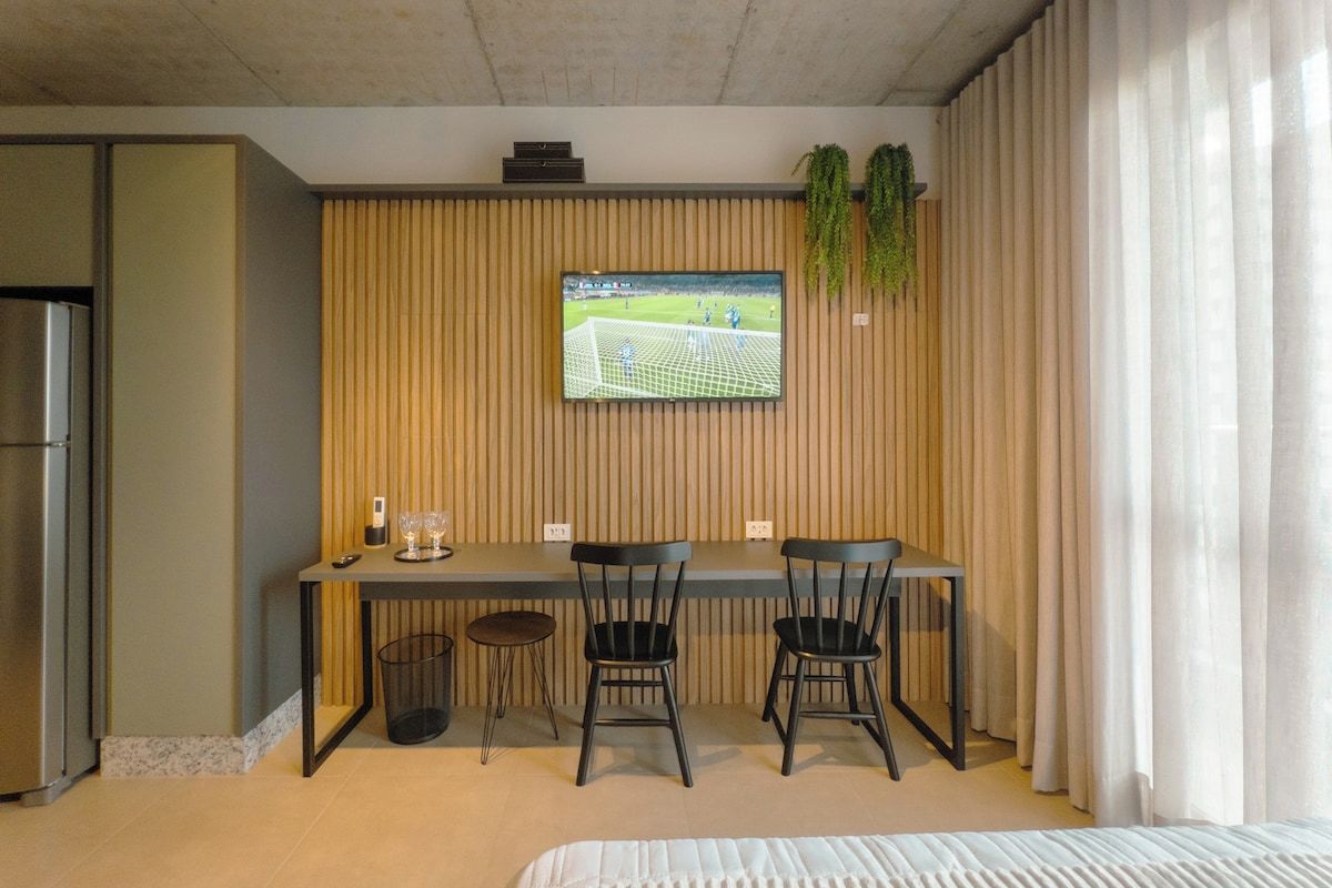 Moderni hotellihuone, jossa on sänky, puiseen seinään asennettu televisio, pieni pöytä tuoleineen ja roskakori.