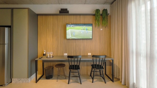 Quarto de hotel moderno com cama, televisão fixada numa parede de madeira, mesa pequena com cadeiras e um cesto de lixo.