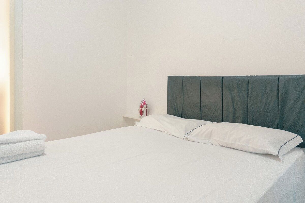 A neatly made bed with white linens and a dark headboard in a simple room with a white wall and a small nightstand with a red object.