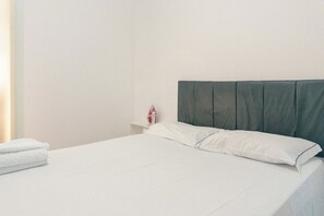 Ein ordentlich bezogenes Bett mit weißen Leinen und einem dunklen Kopfteil in einem schlichten Zimmer mit weißer Wand und einem kleinen Nachttisch, auf dem ein rotes Objekt steht.