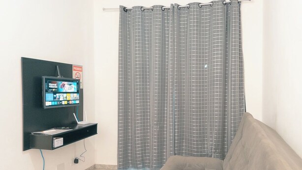 A room with a grey checkered curtain, a mounted television, a sofa, and a small shelf with a device. - Affordable Apartment with AC in Zona 7 - CED103 (Maringá)