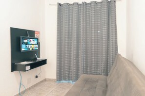 Een kamer met een grijs geruit gordijn, een wandtelevisie, een grijze bank en een zwarte plank met een apparaat.