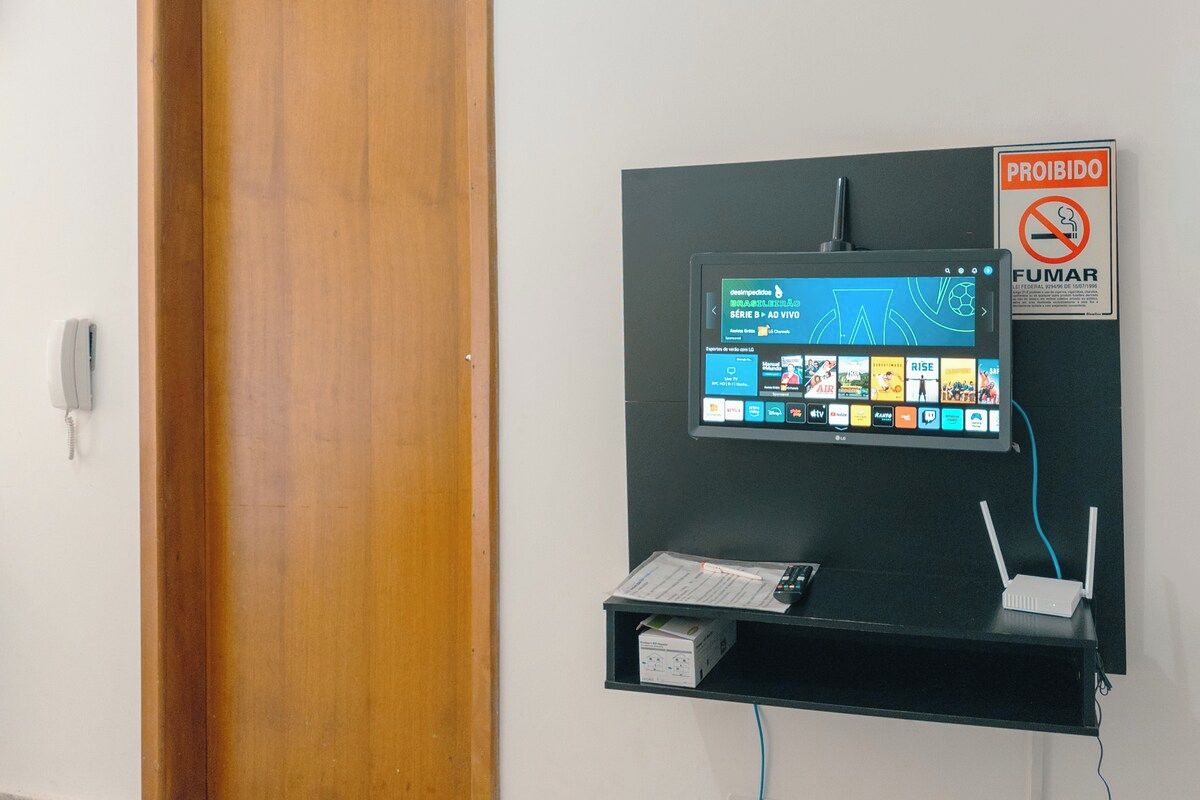 Televisor montado en la pared mostrando una interfaz de servicio de streaming, un letrero de 'PROIBIDO FUMAR' y un estante pequeño con un teléfono y una caja.