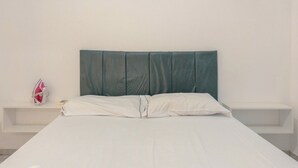 A bed with a headboard, white bedding, and a small shelf with a red object. - Affordable Apartment with AC in Zona 7 - CED103 (Maringá)