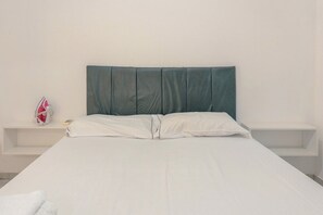 Ein Bett mit Kopfstütze, weißer Bettwäsche und einem kleinen Regal mit einem roten Gegenstand.