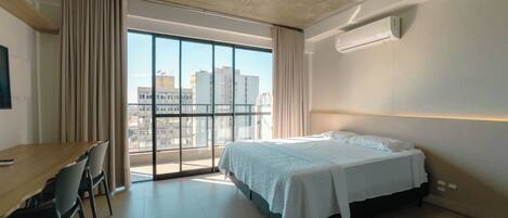 A modern hotel room with a large bed, a desk, and a view of the city through large windows.