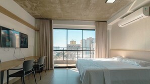 Habitación de hotel con cama, escritorio, sillas, televisión y una ventana grande con vista a la ciudad.