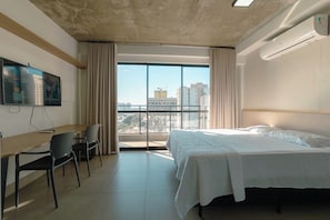 Habitación de hotel con cama, escritorio, sillas, televisión y una ventana grande con vista a la ciudad.