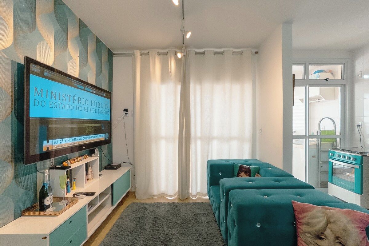 Een woonkamer met een televisie waarop een nieuwskanaal draait, een bank, een keukenhoek en een raam met gordijnen.