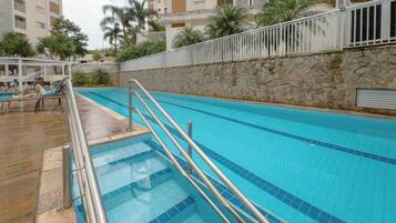 Una piscina con pared de piedra y barandillas de metal, rodeada de palmeras y un edificio al fondo.