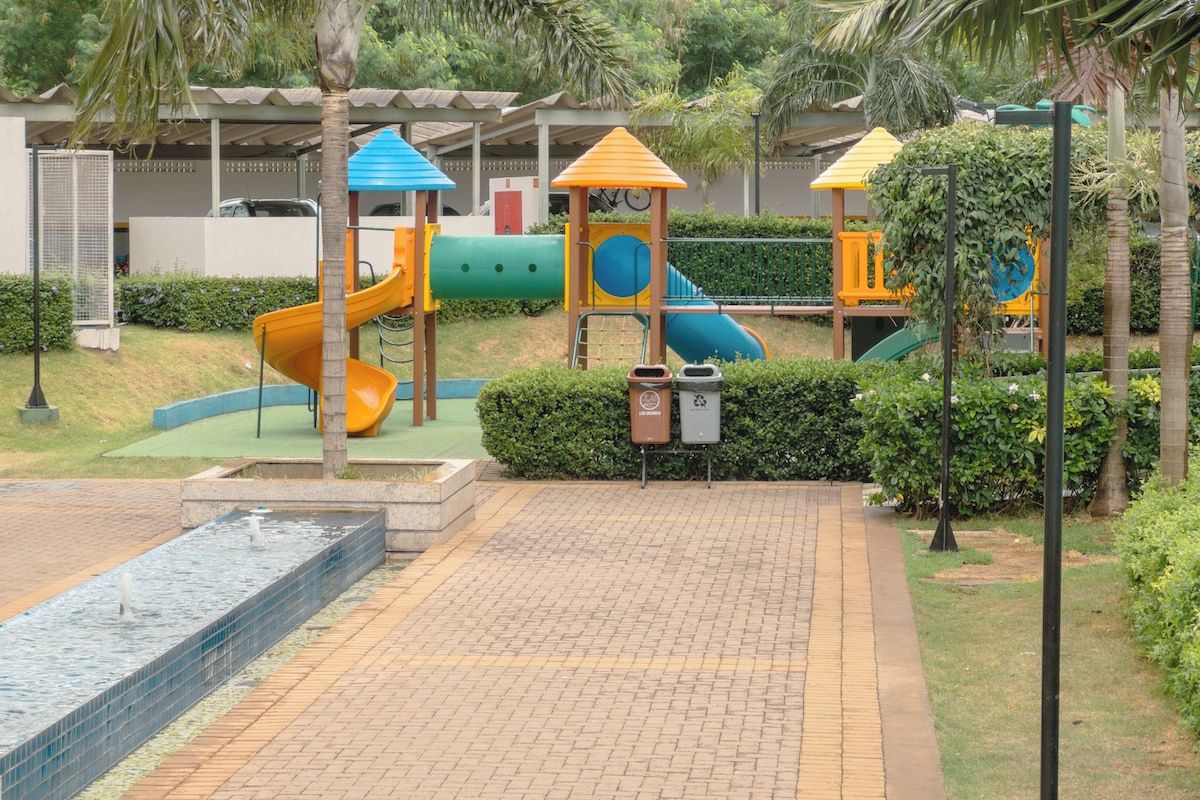 A playground with slides and climbing structures, surrounded by a paved area and a water feature.