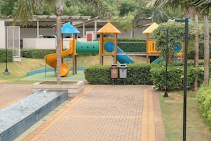 A playground with slides and climbing structures, surrounded by a paved area and a water feature.