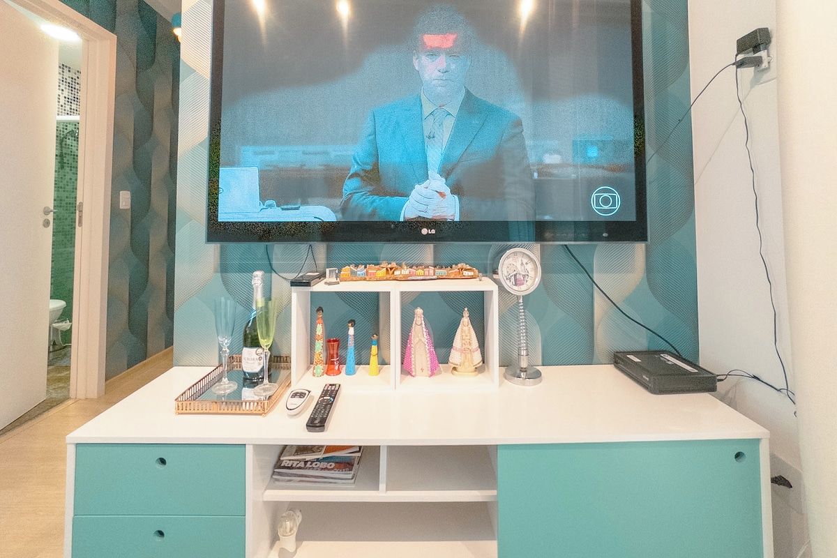 Un televisor montado en la pared, un escritorio blanco con cajones, un estante con varios objetos y un control remoto.