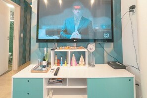 Ein an der Wand befestigter Fernseher, ein weißer Schreibtisch mit Schubladen, ein Regal mit diversen Gegenständen und eine Fernbedienung.