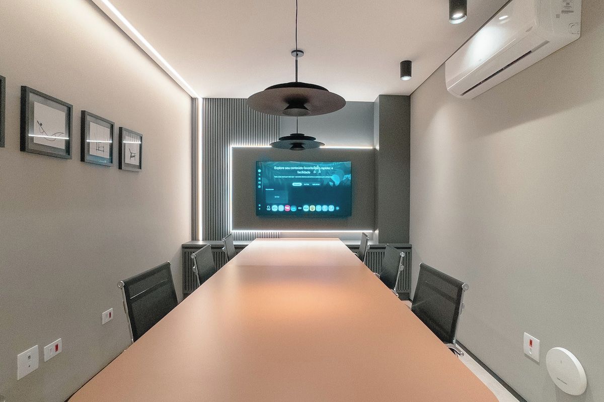 Sala de conferência com uma mesa comprida, cadeiras, um ventilador de teto e uma tela exibindo conteúdo.