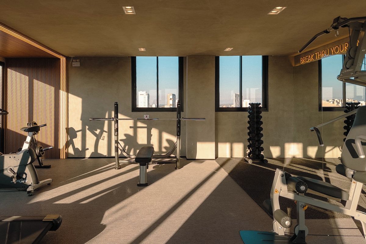 Academia com equipamentos de ginástica, amplas janelas e vista para a paisagem urbana.