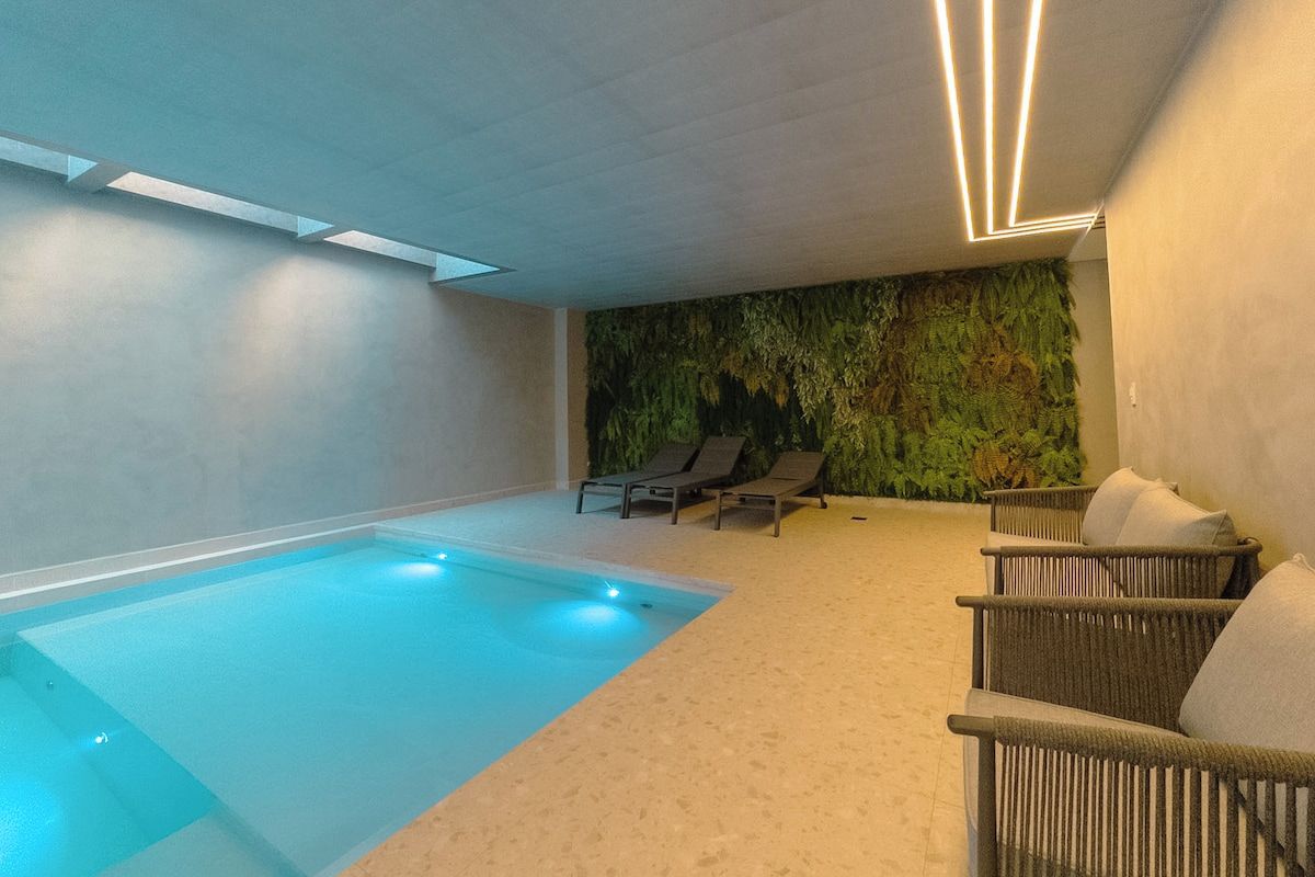 Ein moderner, überdachter Pool mit klar blauem Wasser, umgeben von Liegestühlen und einer strukturierten Wand mit Pflanzen.