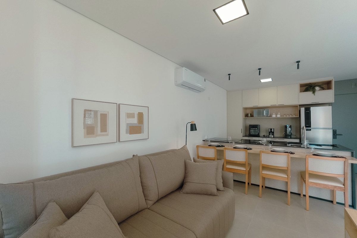 A modern living space with a sofa, a dining area with chairs, a kitchen with cabinets, and an air conditioning unit.