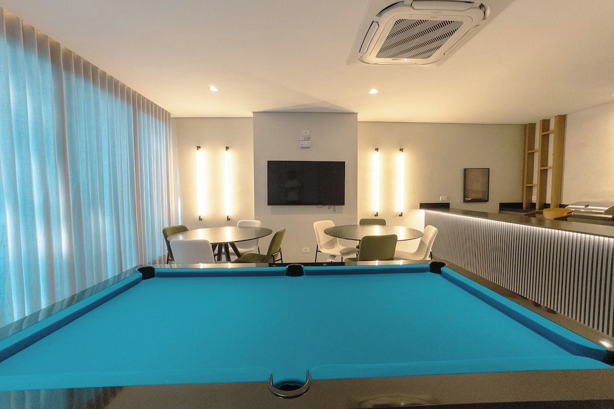 Un espace piscine avec une piscine bleue, des chaises blanches, une table et un téléviseur fixé au mur.
