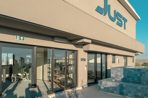 Een gebouw met een ruime glazen ingang, een bord waarop 'JUST' staat en buitentoestellen voor fitness.