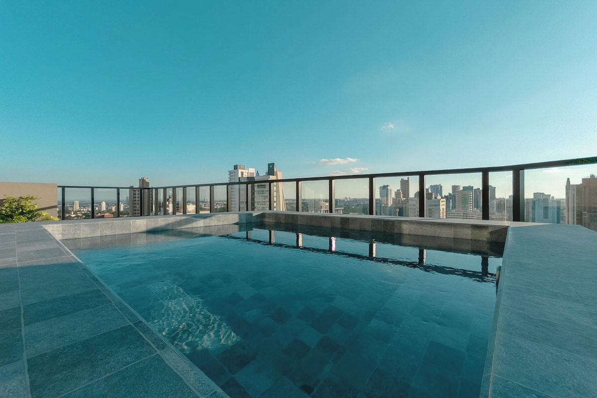 Piscina no terraço com barreira de vidro, vista para o horizonte da cidade e céu claro.