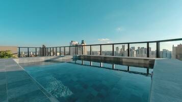 Piscina no terraço com barreira de vidro, vista para o horizonte da cidade e céu claro.