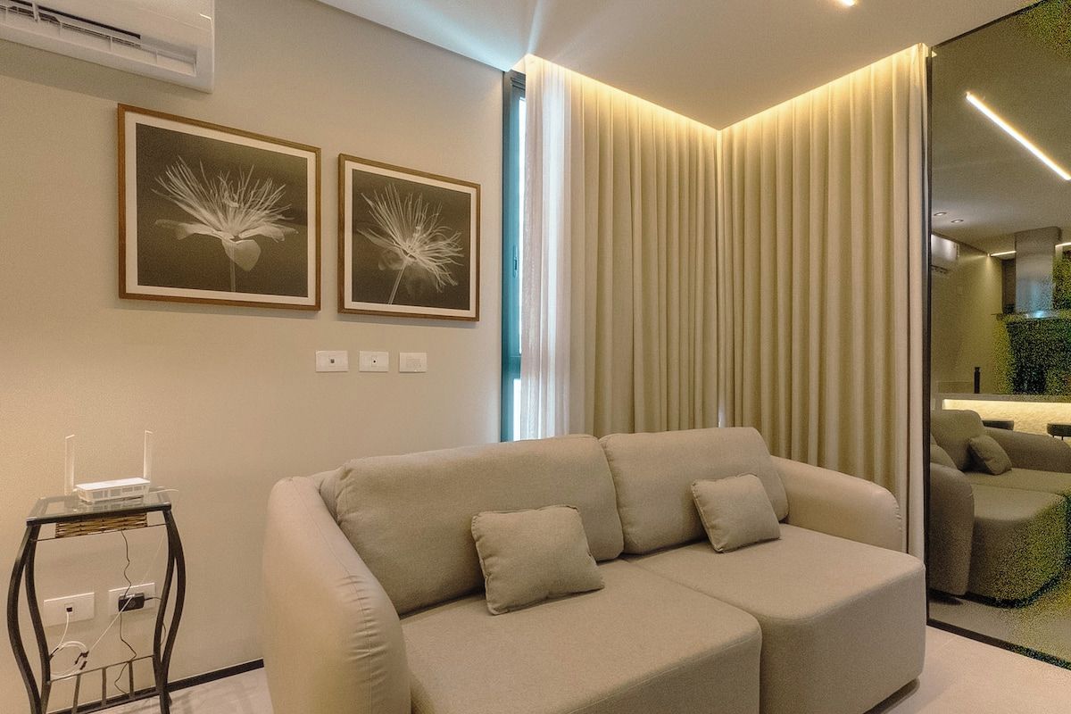 A modern living room with a beige sectional sofa, two framed wall art pieces, a glass side table with a router, and a large window with curtains.