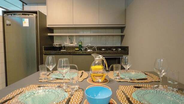 A dining table set with plates, glasses, and cutlery, with a kitchen in the background. - Modern Apartment near UEM with Wi-Fi - LP12 (Maringá)