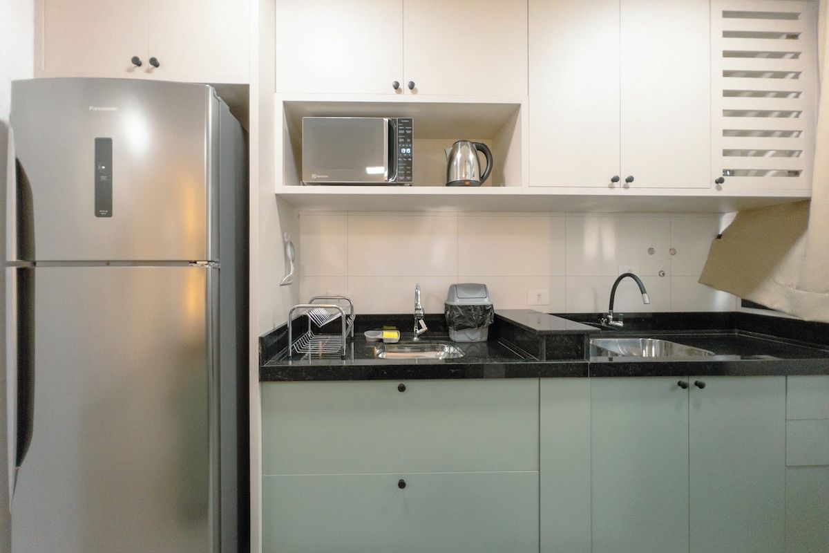 A compact kitchen with a stainless steel refrigerator, a microwave, a kettle, a sink, and a dish rack.
