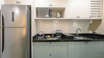 A compact kitchen with a stainless steel refrigerator, a microwave, a kettle, a sink, and a dish rack.