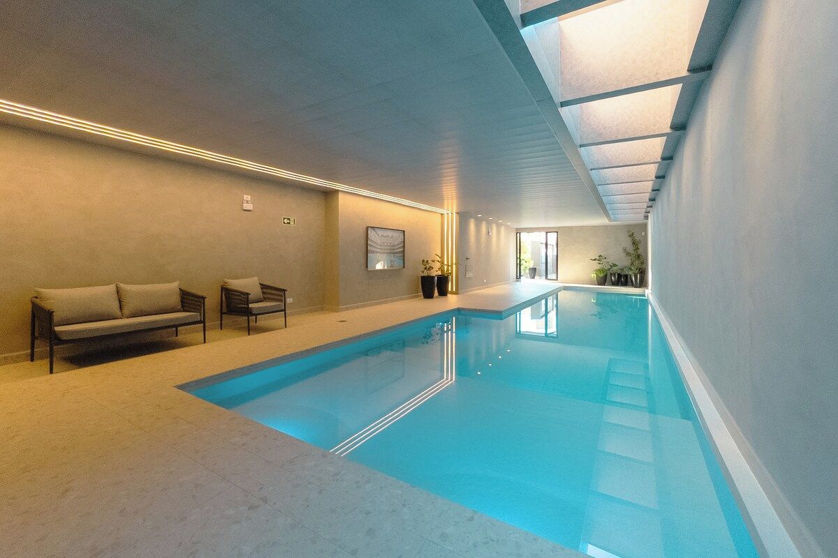 Piscina interna com superfície de água cristalina, teto com claraboias e área de estar com sofás e cadeiras.