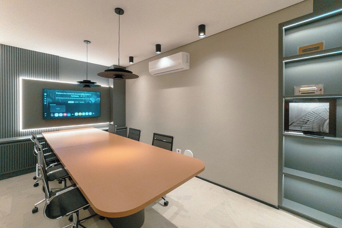 Uma sala de conferências com uma mesa comprida, cadeiras, uma tela de televisão e um aparelho de ar condicionado.