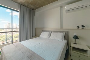 A hotel room with a large bed, a nightstand, a lamp, and a view of the city through a window.