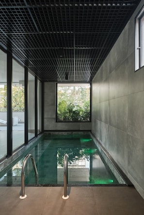 En moderne indendørs swimmingpool med gitterloft, store vinduer og udsigt til grøn vegetation udenfor.
