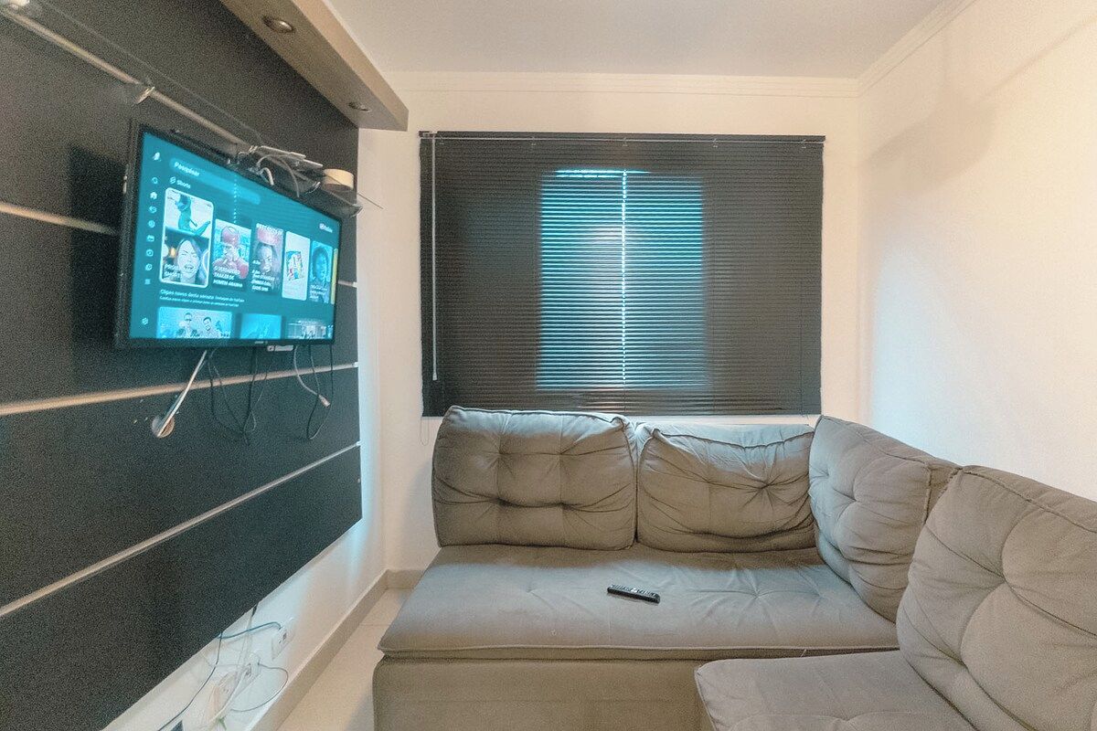 Ein Wohnzimmer mit Flachbildfernseher an der Wand, einer Couch und einem Fenster mit Jalousien.