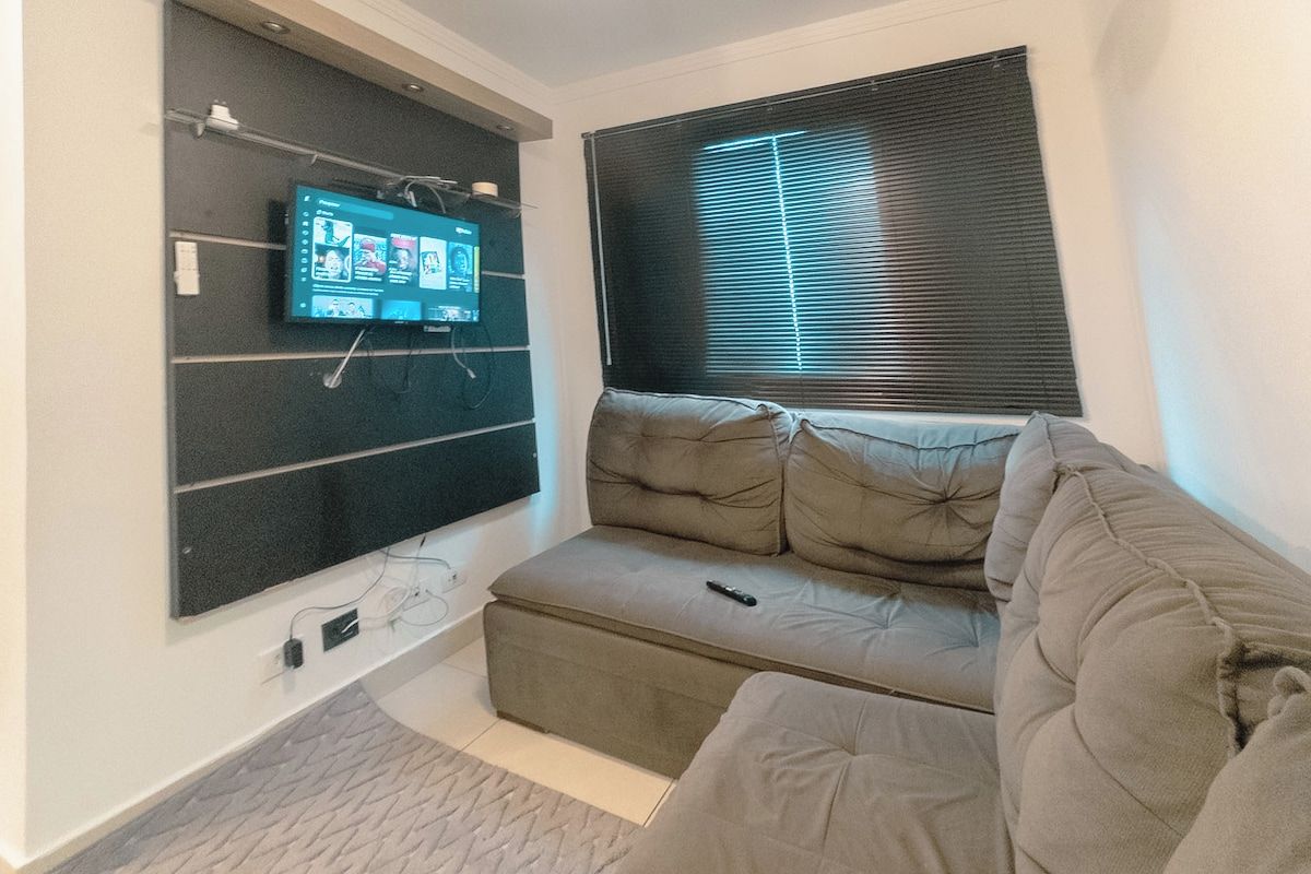 Una sala de estar con un televisor de pantalla plana montado en la pared, un sofá y una ventana con persianas.