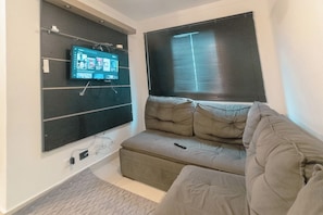 Uma sala de estar com televisão de tela plana na parede, um sofá e uma janela com persianas.