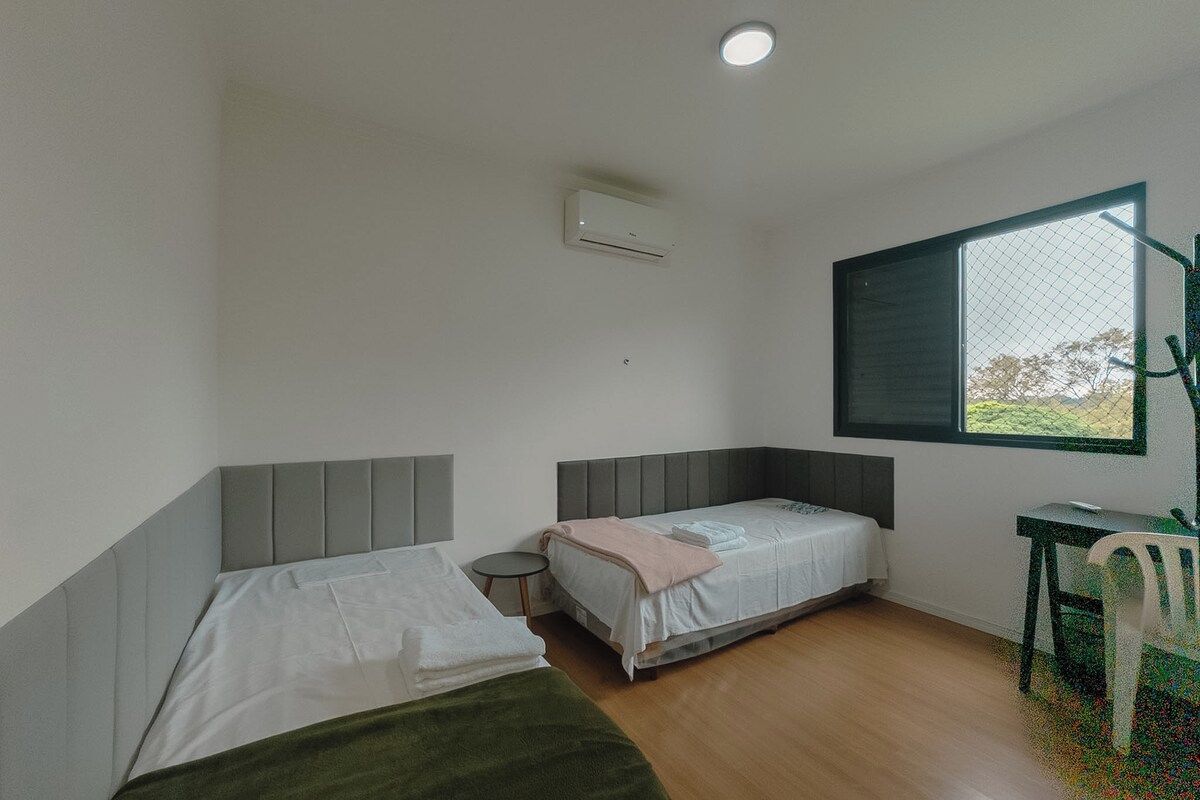 Una habitación con dos camas, una mesita pequeña, una silla, un ventanal y un equipo de aire acondicionado.