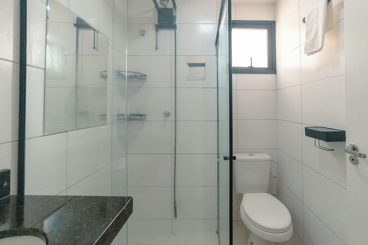 Casa de banho com cabine de chuveiro em vidro, sanita e lavatório com bancada escura.