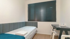 Yhden hengen sänky, jossa on valkoinen patja ja sininen peitto, työpöytä tuolilla sekä ikkuna, jossa on sälekaihtimet.