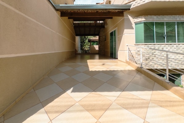 A corridor with tiled flooring, a metal railing on one side, and a covered area with a view of greenery in the distance.