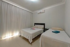 A hotel room with two beds, a white air conditioning unit on the wall, and a window with white curtains.